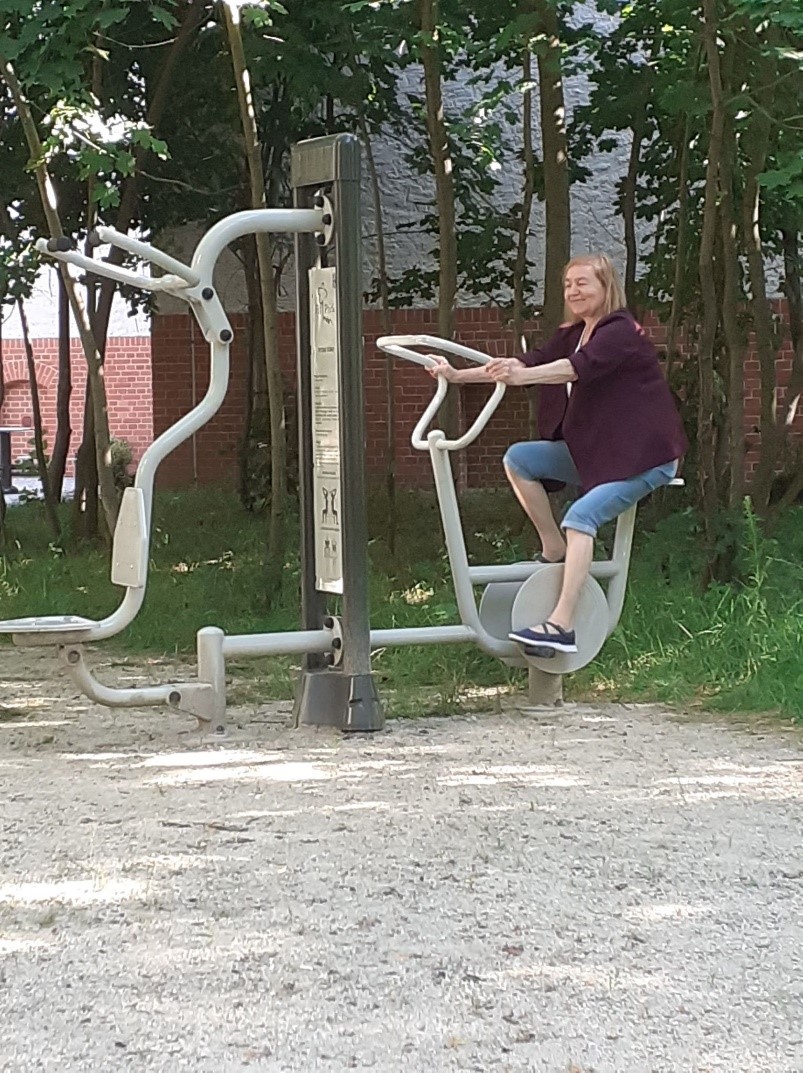 Zdjęcie przedstawia urządzenia do ćwiczeń w ogrodzie. Na jednym z urządzeń ćwiczenie jazdy na rowerze wykonuje klientka DDP. Ubrana jest w bordową marynarkę, niebieskie dżinsowe spodenki do kolan oraz granatowo - białe buty